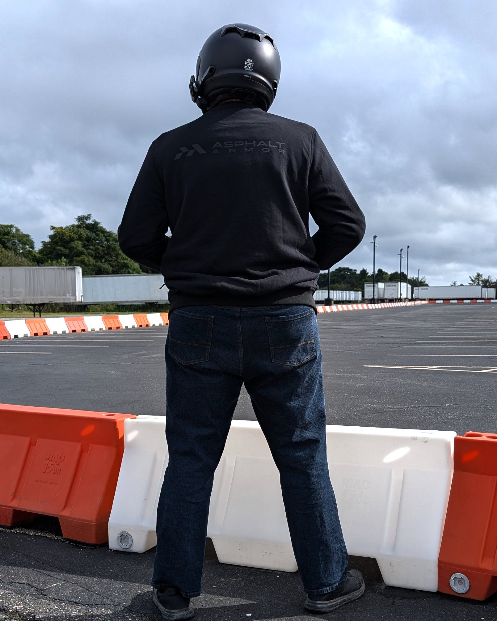 Person wearing a black jacket with a brand logo, standing near orange and white barriers in an open area.